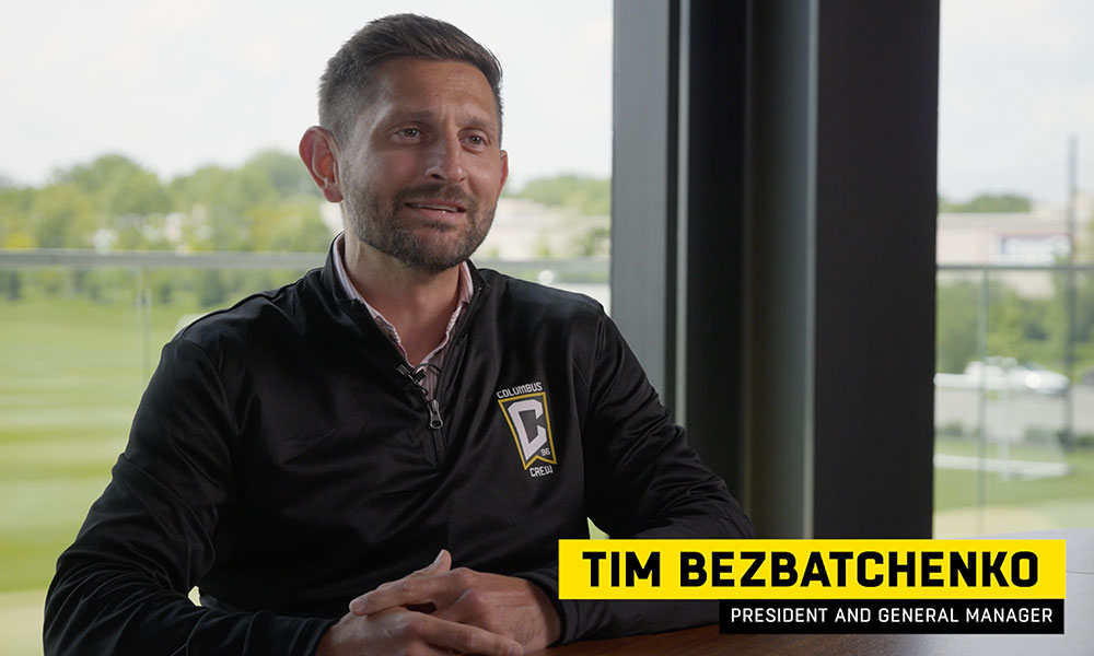 Image of a man wearing a Columbus Crew track jacket being interviewed with the soccer fields in the background