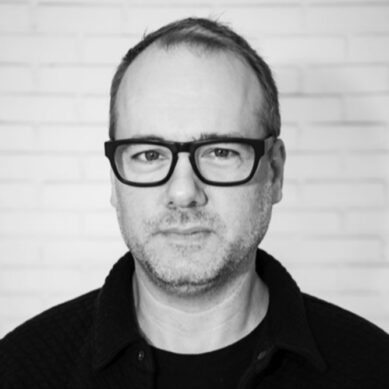 Portrait of a man with glasses against a white brick wall.