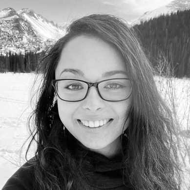 Portrait of a woman with long brown hair and glasses smiling outdoors in the mountains