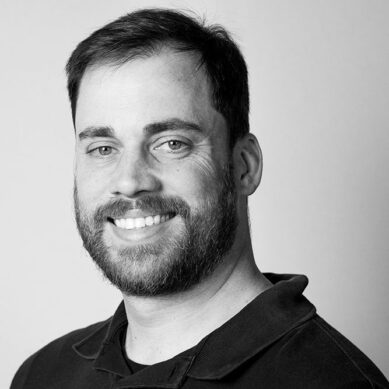 Black and white portrait of a smiling man with a beard, wearing a casual black shirt.